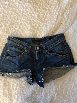 Blå jeansshorts med fransig kant - Säljer ett par blå jeansshorts med rå, fransig kant och klassisk femficksdesign. Shortsen har låg midja och slitna detaljer för en avslappnad look. Perfekta för sommaren och festivaler💖💖💖