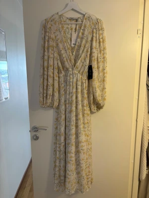 Lång blommig omlottklänning - Långklänning i ljus bas med gult blommönster och v-ringad omlottdesign. Klänningen har långa puffärmar och ett luftigt, svepande fall. Perfekt för sommarens alla tillfällen.