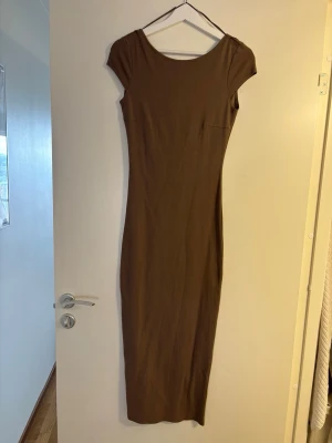 Brun långklänning med kort ärm - En enkel och stilren brun långklänning med rund halsringning och korta ärmar. Klänningen har en figurnära passform och är tillverkad i ett mjukt, stretchigt material som känns bekvämt mot huden. Perfekt för dig som gillar minimalistisk stil. Öppen i ryggen. 