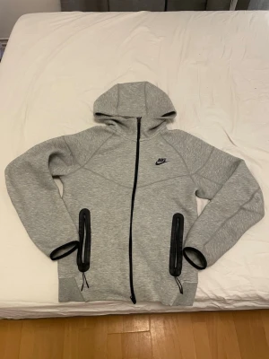 Grått Nike set med huva och dragkedja - Säljer ett grått Nike set med hoodie och matchande byxor. Hoodien har dragkedja, huva och svarta detaljer vid fickorna. Byxorna har resår i midjan, snörning och svarta dragkedjor vid fickorna. Perfekt för en avslappnad och sportig stil.