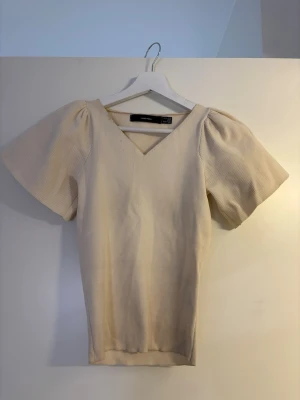Beige v-ringad topp från Vero Moda - Snygg beige topp från Vero Moda med v-ringning och korta, lite puffiga ärmar. Tillverkad i ett mjukt material som känns bekvämt mot huden. Perfekt att styla med jeans eller kjol för en trendig look.