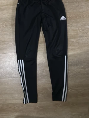 Svarta Adidas träningsbyxor - Säljer ett par svarta träningsbyxor från Adidas med vita ränder längs benen och Adidas-logga på låret. Byxorna har smal passform och är tillverkade i ett stretchigt funktionsmaterial som passar perfekt till träning eller sport.