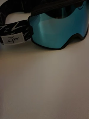Zipe skidglasögon med blått glas - Snygga skidglasögon från Zipe med spegelblått glas och svart/vitt mönstrat band. Perfekta för vinterns skidåkning eller snowboard. Justerbart band för bra passform och bekväm vaddering runt ramen.