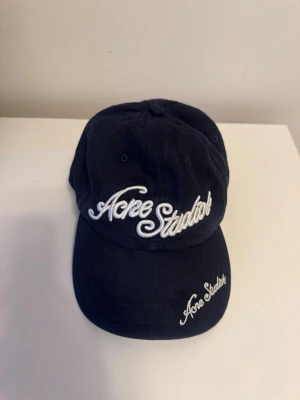 Mörkblå keps från Acne Studios - Snygg mörkblå keps från Acne Studios med vit broderad logga framtill och på skärmen. Klassisk böjd skärm och justerbar passform. Kepsen är i mycket bra skick.