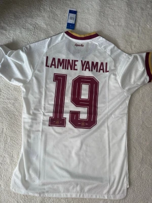 Vit Spanien Lamine Yamal matchtröja - Säljer en vit Spanien fotbollströja från Adidas med Lamine Yamal och nummer 19 på ryggen. Tröjan har vinröda detaljer, gula inslag vid kragen och klassiska Adidas-ränder på axlarna. Tillverkad i lätt och ventilerande material, perfekt för match eller träning.