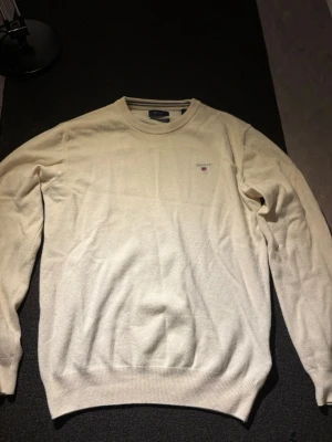 Stickad tröja från Gant - Snygg beige stickad tröja från Gant med rund halsringning och diskret broderad logga på bröstet. Tröjan har långa ärmar och ribbade muddar vid ärmslut och nederkant. Perfekt för en clean och klassisk stil.