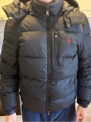 Svart pufferjacka från Polo Ralph Lauren - Säljer en svart pufferjacka från Polo Ralph Lauren med röd logga på bröstet. Jackan har huva, hög krage, dragkedja framtill och en bröstficka med dragkedja. Perfekt för kalla vinterdagar och riktigt skön att bära.
