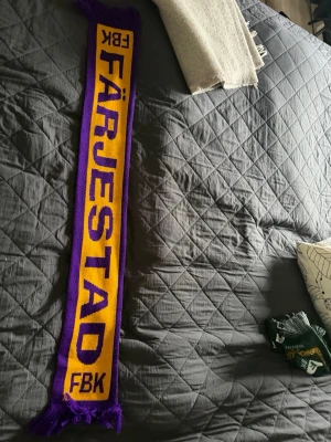 Färjestad supporterscarf lila/gul - Supporterscarf med Färjestad BK-tryck i lila och gult. Klassisk rektangulär form med fransar i ändarna. Perfekt accessoar för att visa ditt lagstöd på matchen eller i vardagen.