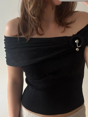 Svart offshoulder topp med brosch - Trendig svart offshoulder topp med ribbad kropp och draperad axelparti. Snygg detalj med brosch och pärla på ena axeln. Perfekt för dig som vill sticka ut med en unik och stilren look.