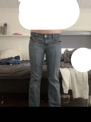 Blå bootcut jeans med fickdetaljer - Säljer ett par blå bootcut jeans med dubbla knappar i midjan och stora fyrkantiga framfickor. Jeansen har markerad söm längs benen och breda hällor för bälte. Klassisk tvättad denim-look och raka ben med lätt utsvängning.