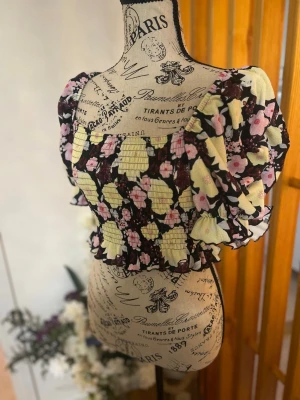Blommig croppad blus med volang - Supersöt croppad blus med blommigt mönster i svart, gul och rosa. Puffiga korta ärmar och volangdetaljer vid ärmslut och nederkant. Perfekt för dig som vill ha en trendig och färgglad topp med romantiska vibbar.