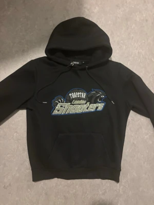 Trapstar Shooters hoodie - Säljer en svart hoodie från Trapstar med trycket 'London Shooters' i blått, vitt och silver på bröstet. Hoodien har huva med dragsko och en stor magficka. Perfekt för dig som gillar streetwear och vill sticka ut med en unik design.