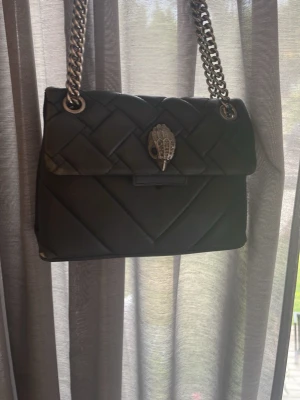 Svart quiltad handväska Kurt Geiger - Snygg svart quiltad handväska från Kurt Geiger med silverfärgad kedjerem och ikonisk örndetalj på locket. Väskan är väl andvänd, köpt ny för ungefär 2500kr