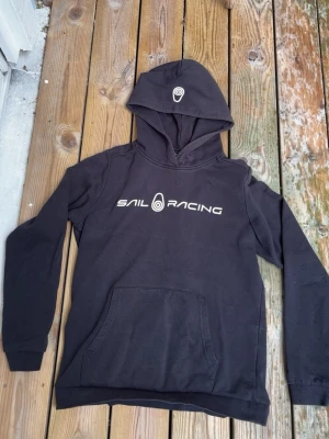 Svart hoodie från Sail Racing - Svart hoodie från Sail Racing med vit logga och text på bröstet samt logga på huvan. Klassisk känguruficka framtill och ribbade muddar vid ärmslut och nederkant. Perfekt för en avslappnad och sportig stil.