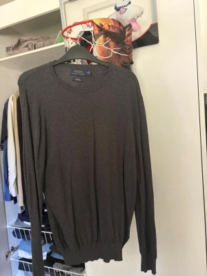 Grå stickad tröja från Polo Ralph Lauren - Mjuk och stilren grå stickad tröja från Polo Ralph Lauren med lila broderad logga på bröstet. Tröjan har rund halsringning, långa ärmar och ribbade muddar vid ärmslut och nederkant. Perfekt till lager på lager och enkel att matcha.
