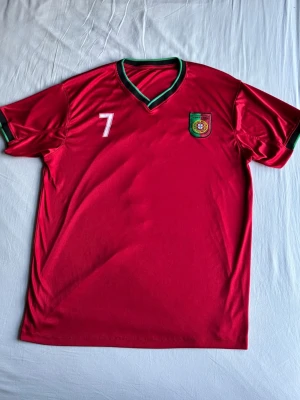 Ronaldo fotbolls tröja  - Säljer en röd Portugal fotbollströja med gröna detaljer, nummer 7 och Ronaldo på ryggen. Tröjan har V-ringning och korta ärmar, samt Portugals landslagsemblem på bröstet. Perfekt för dig som vill stötta landslaget eller bära en ikonisk spelare. Storlek xxl men sitter som L.