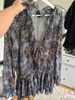 Mönstrad kortklänning från Monki - Säljer en kortklänning från Monki med blandat mönster i grå, brun och blå toner. Klänningen har volangdetaljer, snörning framtill och långa ärmar. Materialet känns lätt och flowy, perfekt för dig som gillar en unik och bohemisk stil.