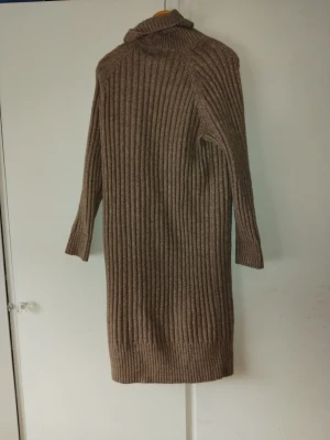 Brun ribbad stickad långklänning H&M - Mysig brun ribbad långklänning från H&M i stickat material. Klänningen har polokrage och långa ärmar, perfekt för kyligare dagar. Enkel och stilren design som är lätt att matcha med olika accessoarer.