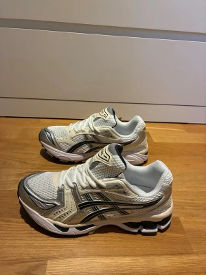 Vita och silver ASICS sneakers - Snygga sneakers från ASICS i vitt och silver med mesh- och syntetpaneler. Skorna har en sportig vibe med svarta detaljer och klassisk ASICS-logga på sidan. Perfekta för dig som gillar retrostil och bekväma skor med bra dämpning.