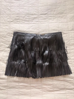Svart partykortkjol med fransar - Säljer en svart kortkjol med fransar som ger en cool och edgy look. Kjolen är tillverkad i ett syntetmaterial som liknar skinn. Perfekt för en fest eller party.