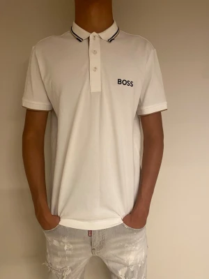 Vit pikétröja från BOSS - Snygg vit pikétröja från BOSS med svart logga på bröstet och krage med tunna svarta ränder. Regular fit med stretchigt material för extra komfort. Klassisk knappslå med tre knappar och korta ärmar. Perfekt till jeans eller chinos.