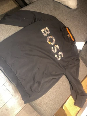 Svart sweatshirt med BOSS-tryck - Svart sweatshirt med stort BOSS-tryck i mönstrade bokstäver på bröstet. Klassisk rund halsringning och ribbade muddar vid ärmslut och nederkant. Perfekt för en avslappnad och trendig look.