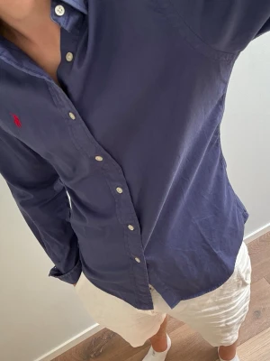 Marinblå skjorta från Ralph Lauren - Säljer en marinblå skjorta från Ralph Lauren med klassisk krage och knappar framtill. Skjortan har lång ärm och den ikoniska röda logotypen broderad på bröstet. Tillverkad i bomull för en skön och stilren look.