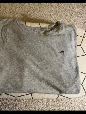 Grå t-shirt från Gant - Klassisk grå t-shirt från Gant med liten broderad logga på bröstet. T-shirten har rund halsringning och korta ärmar. Tillverkad i mjuk bomull som känns skön mot huden. Perfekt basplagg till garderoben.