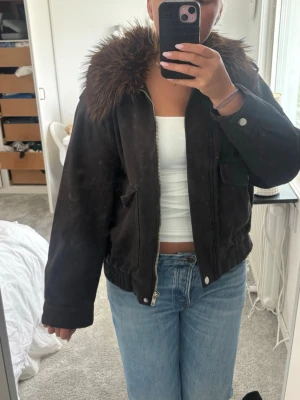 Svart jacka med fuskpäls-krage - Snygg svart jacka med stor brun fuskpäls runt kragen. Jackan har dragkedja framtill, fickor med lock och resår i midjan. Perfekt för dig som vill ha en cool och trendig look med lite extra edge.