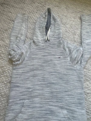 Gråmelerad hoodie från Hollister - Säljer en gråmelerad hoodie från Hollister med vit dragsko i huvan och klassisk Hollister-logga på bröstet. Hoodien har känguruficka framtill och är tillverkad i mjukt, stickat material. Perfekt för chill dagar och snygg till jeans. Skickar samma dag eller dagen efter🚚 rök och djurfritt hem!