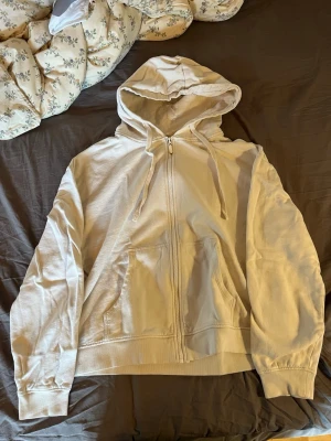 Beige/ vit zip hoodie med huva - Säljer en beige/vit hoodie med dragkedja och huva. Hoodien har två stora fickor framtill och justerbara snören i huvan. Passar perfekt till chill dagar och är gjord i mjukt bomullsliknande material. Enkel och clean design utan synligt märke.