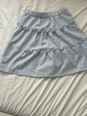Blommig blå volangkjol - Söt blå kjol med små vita blommor och volangdetaljer. Kjolen har resår i midjan och är i en kort modell, perfekt för varmare dagar. Materialet är mjukt och lätt, troligen bomull eller polyester. Passar dig som gillar romantisk och avslappnad stil. Kjolen är väldigt stretchig i midjan. 