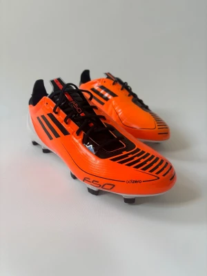 Adidas F50 adizero orange fotbollsskor - Säljer ett par grymma Adidas F50 adizero fotbollsskor i en riktigt snygg orange färg med svarta detaljer och vita sulor. Skorna har klassiska svarta ränder på sidorna, snörning och dobbar för bra grepp på planen. Perfekta för dig som vill sticka ut på matchen! Helt nya.
