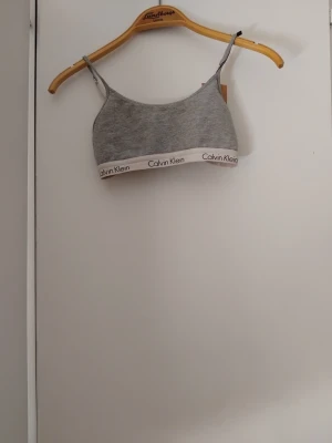 Grå bralette från Calvin Klein - Säljer en stilren grå bralette från Calvin Klein med tunna, justerbara axelband och elastiskt band med logga i vitt. Mjuk och bekväm modell utan bygel, perfekt för en avslappnad look. Materialet är bomullsblandning för extra komfort.
