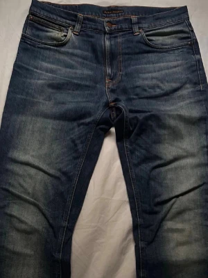 Blå jeans från Nudie Jeans - Säljer ett par klassiska blå jeans från Nudie Jeans med snygg slitning och kontrastsömmar. Jeansen har fem fickor, orange broderi på bakfickorna och normal passform. Perfekta för dig som gillar en avslappnad men stilren look.