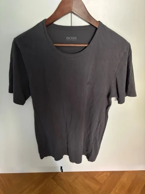 Mörkgrå t-shirt från BOSS - Snygg mörkgrå t-shirt från BOSS med diskret logga broderad på bröstet. Klassisk rund halsringning och korta ärmar. Tillverkad i mjukt material som känns skönt mot huden. Perfekt basplagg för en stilren och enkel look.