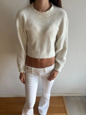 Vit croppad stickad tröja - Säljer en vit croppad stickad tröja med rund hals och långa ärmar. Tröjan har en mjuk, fluffig känsla och ribbade muddar vid ärmslut och nederkant. Perfekt för lager på lager och en clean look.