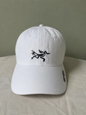 Arcteryx keps - Snygg vit keps med böjd skärm och svart broderad logga framtill. Justerbart spänne bak i metall för perfekt passform. Tillverkad i bomullstyg som känns lätt och skönt på huvudet. Perfekt accessoar för en clean och stilren look.