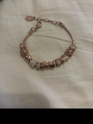 Roséguld armband med pärlor och hjärtan - Snyggt armband i roséguld med flera detaljerade pärlor och hjärtformade berlocker. Armbandet har en kedja med karbinlås och glittrande inslag på vissa pärlor. Perfekt accessoar för att lyfta din stil.