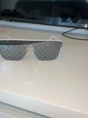 Solglasögon  - Säljer ett par riktigt snygga solglasögon med spegelblanka, silvriga glas och monogrammönster över hela linsen. Sköldformad design med vita skalmar ger en futuristisk vibe. Perfekt accessoar för dig som vill sticka ut.