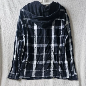 Hollister rutig overshirt med huva - Säljer en svart och vit rutig overshirt med huva. Skjortjackan har långa ärmar, knappar framtill och en avslappnad passform. Perfekt för lager på lager och en cool streetstyle-look.