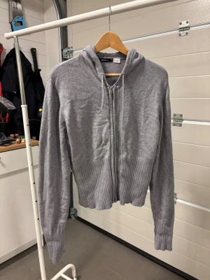 Grå finstickad zip hoodie med ribbade detaljer - Grå finstickad hoodie med dragkedja framtill och huva med dragsko. Tröjan har ribbade muddar vid ärmslut och nederkant samt en croppad passform. Tillverkad i mjukt stickat material som känns skönt mot huden. Perfekt för en avslappnad och stilren look.