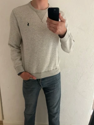 Ljusgrå sweatshirt från Polo Ralph Lauren - Ljusgrå sweatshirt i mjuk bomullsblandning från Polo Ralph Lauren. Klassisk rund halsringning, ribbade muddar och diskret broderad logga på bröstet. Perfekt för en avslappnad och stilren look.
