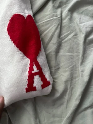 Vit stickad tröja med rött hjärta - Vit stickad tröja med ett stort rött hjärta och bokstaven A på ärmen. Tröjan har rund halsringning, långa ärmar och ribbstickade muddar vid ärmslut och nederkant. Perfekt för dig som gillar stilrena plagg med en twist.