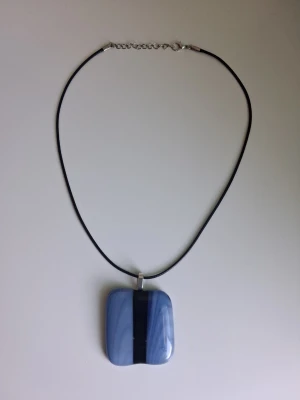 Halsband med blått/svart glas-hänge handgjort glasfusing - Snyggt halsband med ett fyrkantigt blått glas-hänge som har en svart rand i mitten. Hänget har mjuka kanter och sitter på ett tunt svart band med silverfärgat spänne. Perfekt för dig som gillar unika accessoarer och vill sticka ut lite extra. Skapat med hjälp av en hot pot. Storlek ca 4.5cm x 4.5cm 