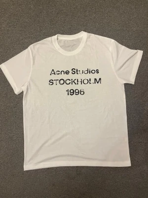 Vit Acne Studios t-shirt Stockholm - Vit t-shirt från Acne Studios med svart text 'STOCKHOLM 1996' på bröstet. Klassisk rund hals och korta ärmar. Perfekt basic-plagg med stilren design och tryck framtill.