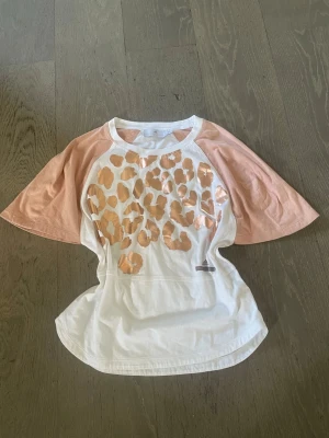 Vit och rosa t-shirt med leopardmönster - Snygg t-shirt med vita och ljusrosa toner, breda raglanärmar och glansigt leopardmönster i kopparfärg framtill. Perfekt för dig som vill sticka ut med en unik och trendig look. Tillverkad i mjuk bomull för skön känsla hela dagen.