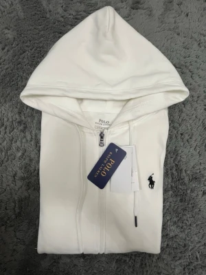 Vit hoodie från Polo Ralph Lauren - Snygg vit hoodie med dragkedja från Polo Ralph Lauren. Klassisk design med huva och justerbara snören, samt den ikoniska svarta polospelaren broderad på bröstet. Tillverkad i mjukt material som känns skönt mot huden. Perfekt för en clean och stilren look.