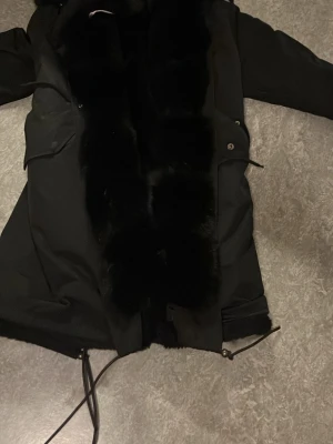 Svart kappa med fuskpälsdetaljer - Snygg svart kappa med stora fickor och dragkedja samt knappar framtill. Kappan har en bred krage och detaljer i svart fuskpäls längs hela framsidan och runt huvan. Perfekt för kalla dagar och ger en lyxig känsla till din outfit.