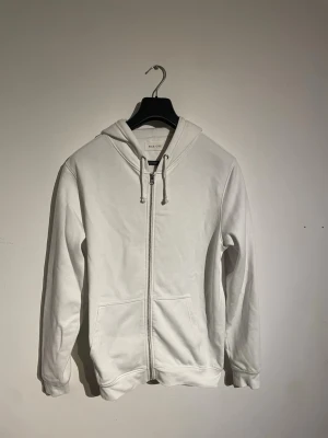 Vit zip hoodie med huva - Snygg vit hoodie med dragkedja framtill och två fickor. Klassisk modell med huva och justerbara snören. Perfekt för en clean och enkel stil. Tillverkad i mjukt material som känns skönt mot huden. Nypris 800kr